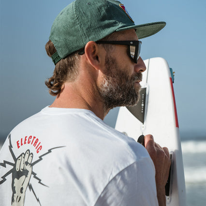 electric brand tee shirt in white worn by model with surfboard. hand with bolt electric brigade
